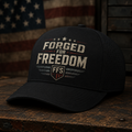Black cap with 'Forged for Freedom' text on a wooden surface with an American flag in the background